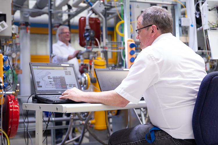 Person überwacht eine technische Prüfanlage über einen Laptop in einer Labor- und Entwicklungsumgebung.